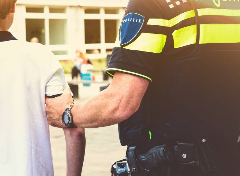 Een politieagent in uniform heeft een kind in wit t-shirt bij  ferm bij de bovenarm vast op een schooplein. Op de achtergrond de grijze stoeptegels van een schoolplein en onscherp een deel van het schoolgebouw.