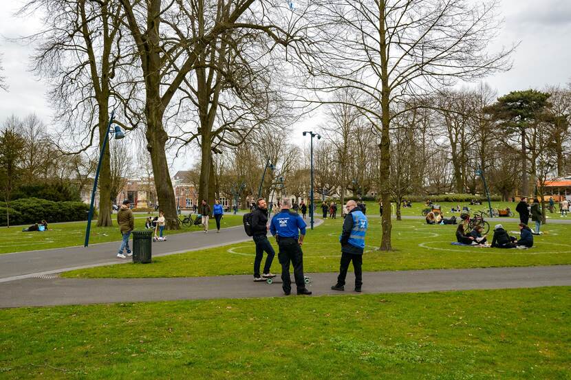 Twee BOA's praten met een burger in een park tijdens hun toezicht op de coronamaatregelen. In het gras van het park zijn witte cirkels te zien waar groepjes mensen in zitten.