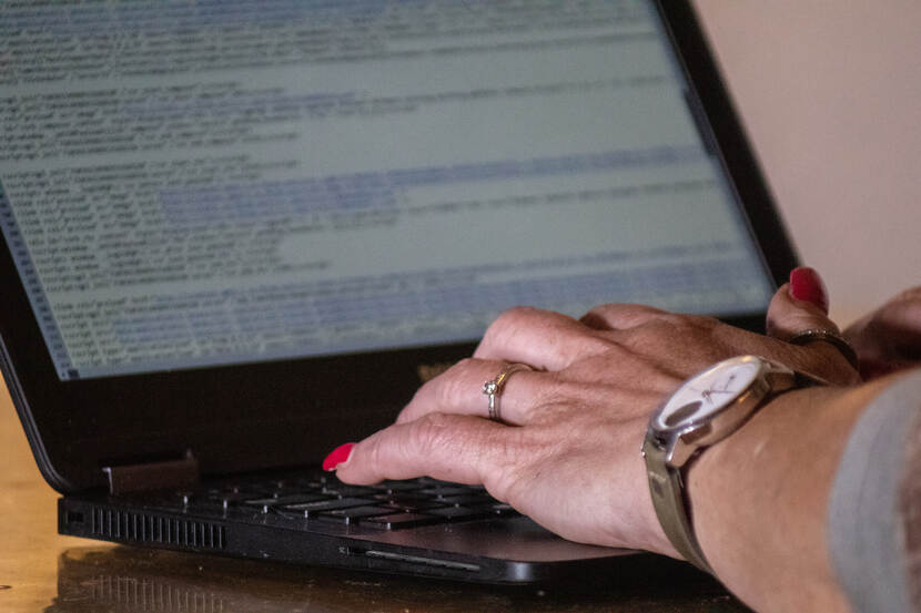 Handen met ringen en rode nagellak boven een toetsenbord van een laptop. Op het scherm is onscherp html-code waarneembaar.