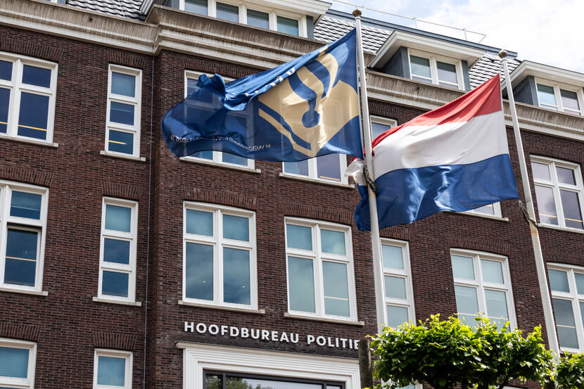 Gebouw met op de gevel  'Hoofdbureau Politie' en op de voorgrond twee wapperende vlaggen. De linker vlag bevat het politie logo en de rechter is de Nederlandse vlag.