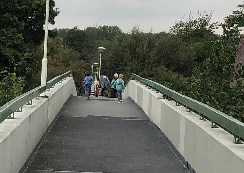 Een voetgangersbrug in een gebied met bomen, gezien over het voetpad. Aan andere kant van de brug, aan de overkant: een groepje kinderen op de rug bezien. Zij wandelen in de richting van de bomen die ook achter de brug te zien zijn.