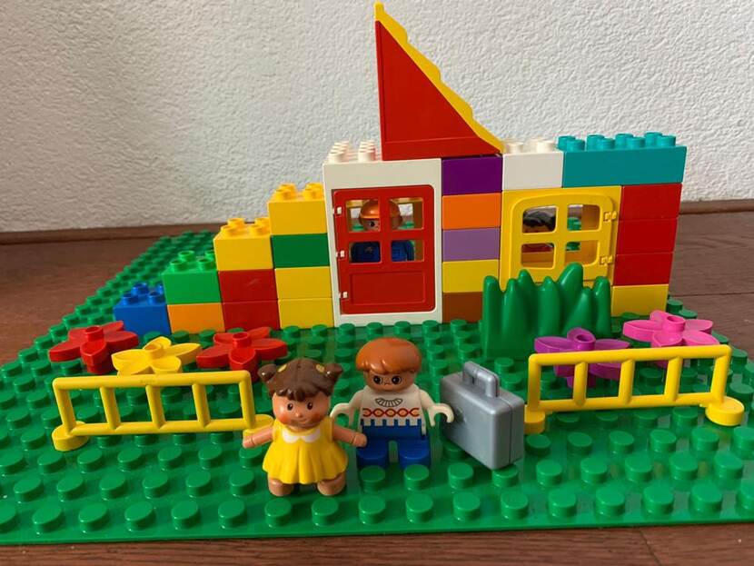 Legohuisje met daarvoor twee poppetjes met een koffer en binnen een poppetje achter de dichte deur.