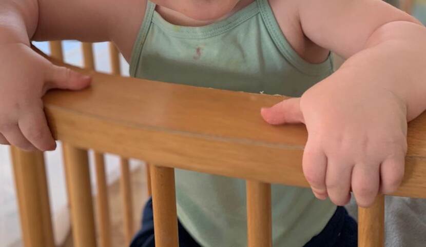 Een klein kind in een groen hemdje leunt met de handen op de leuning van een houten stoel.