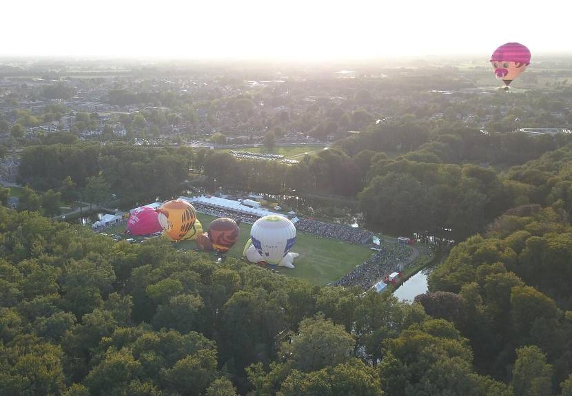 Luchtfoto van ballonfiesta in Barneveld met daarop  circa 8 luchtballonnen nog op de grond en één roze al hoog boven de bomen