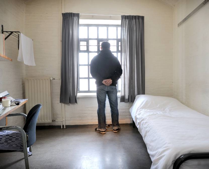 Een gedetineerde staat in zijn cel op de rug bezien midden voor het raam met tralies naar buiten te kijken. Hij heeft zijn handen op zijn rug. Links van hem een tafeltje tegen de ene muur en rechts van hem een opgemaakt bed tegen de andere muur.
